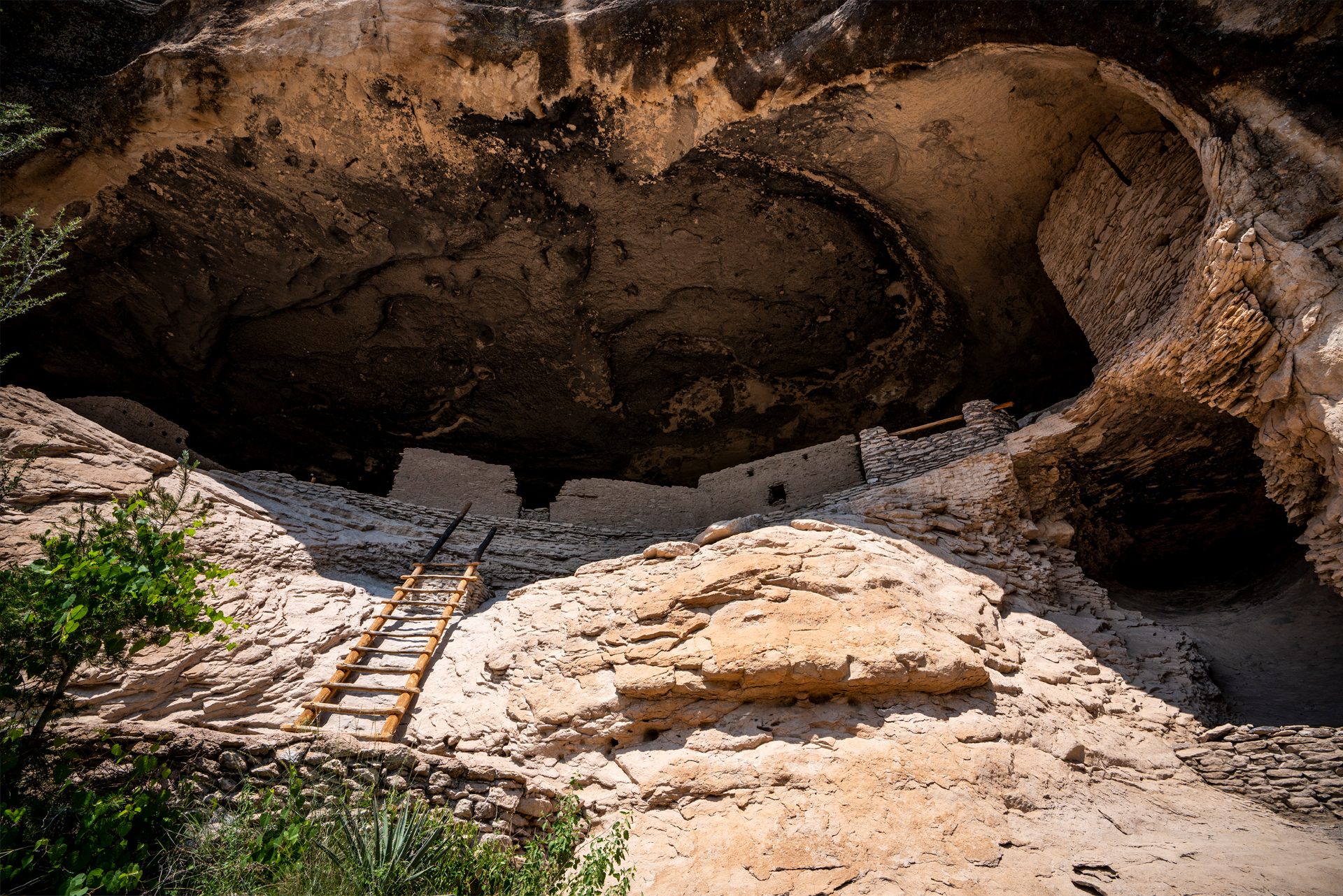 A real photograph of the Gila Cliff Dwellings cave rooms with stone walls and wooden ladders inside a shaded alcove, sunlight spilling in from the canyon opening