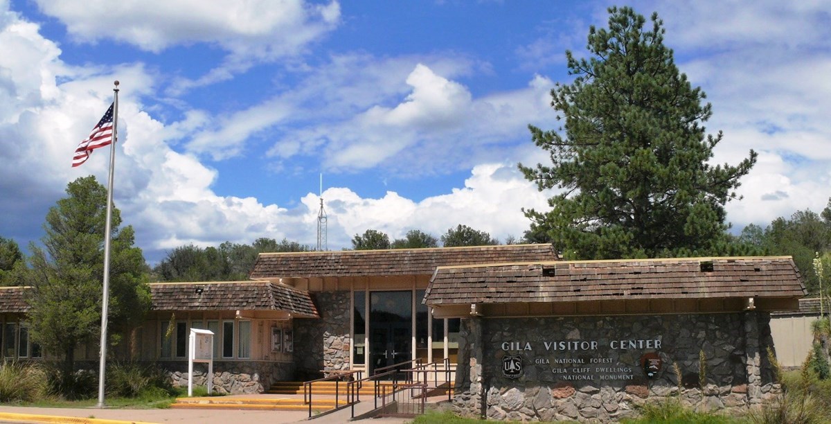 A real photograph of the Gila Cliff Dwellings visitor center exterior with rustic architecture, trees around it, and clear sky in the Gila region