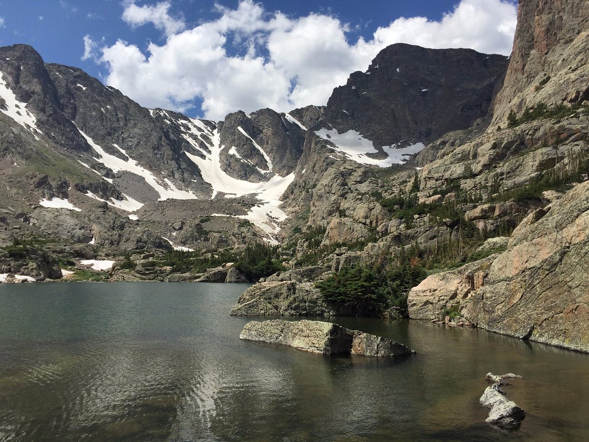 A real photograph of the Glacier Gorge Trailhead sign and nearby parking area in Rocky Mountain National Park at dawn, with headlamps and hikers gearing up