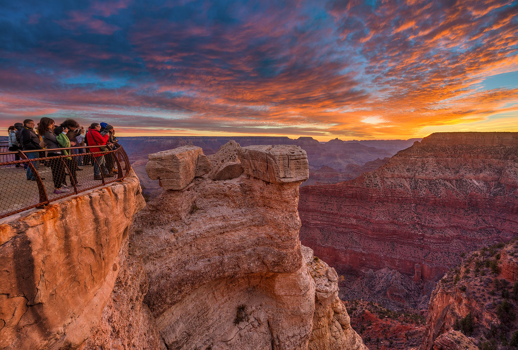 A real photograph of the Grand Canyon South Rim at sunrise from a popular overlook, layered canyon ridges glowing orange and pink with visitors standing near the stone wall