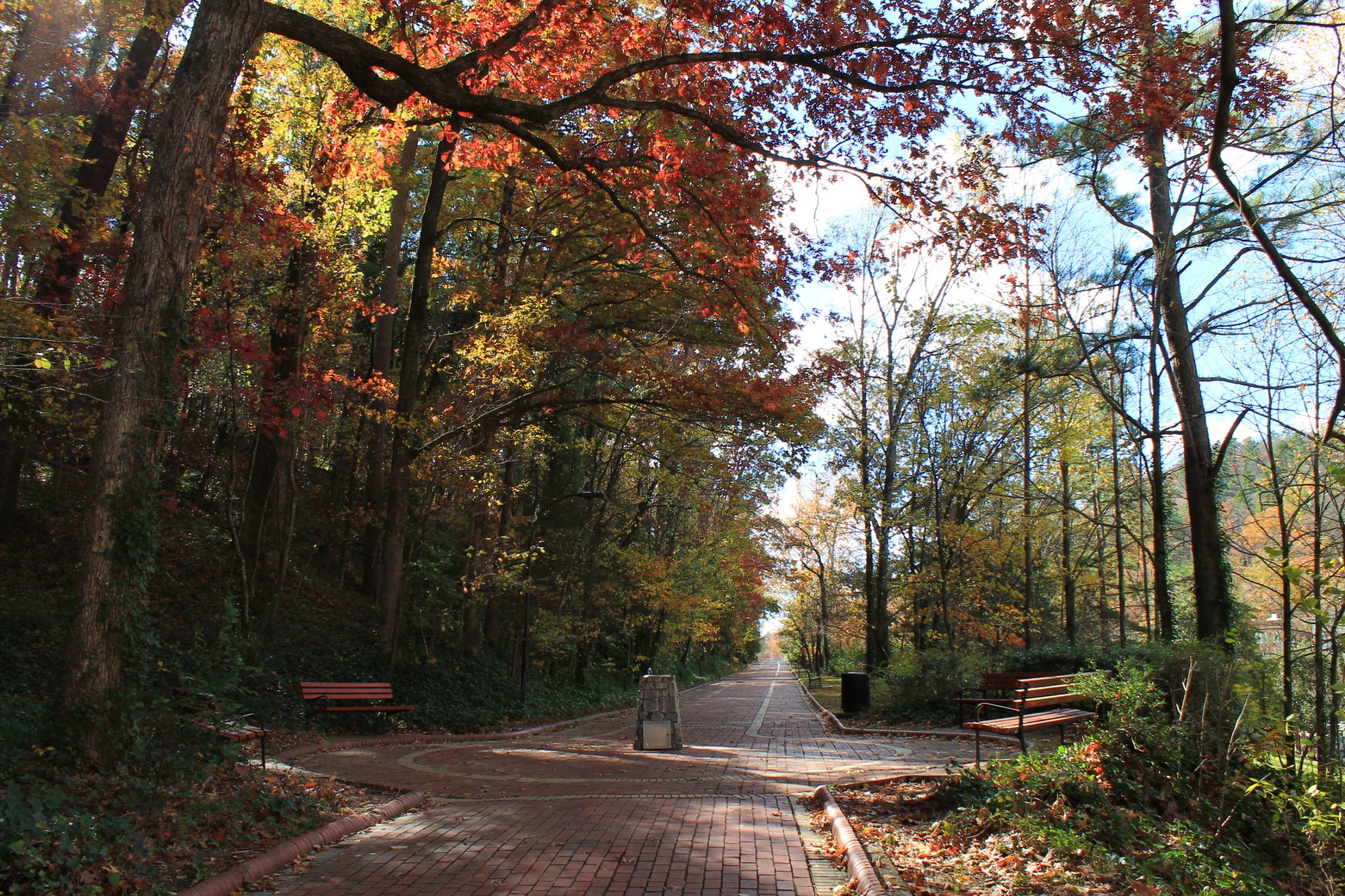 A real photograph of the Grand Promenade walkway above Bathhouse Row in Hot Springs National Park, with a wide brick path, stone walls, and trees on a calm morning