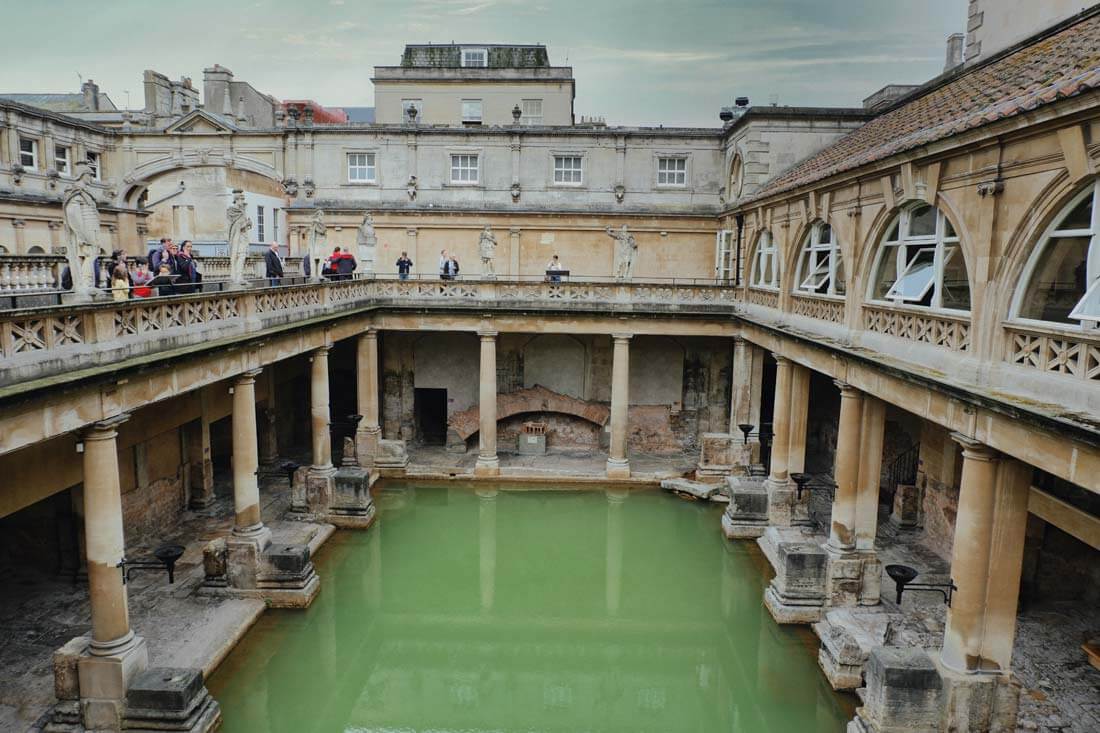 A real photograph of the Great Bath at the Roman Baths in Bath with green steaming water, ancient stone columns and statues around the pool, visitors standing along the edge, indoor-outdoor museum lighting