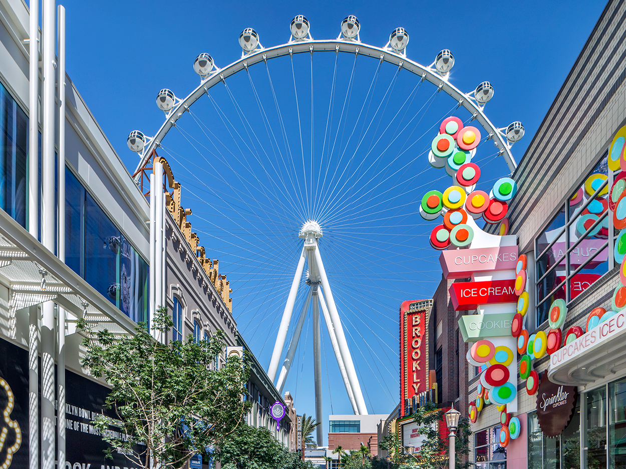A real photograph of the High Roller observation wheel in Las Vegas during the day, with clear blue sky and the surrounding LINQ promenade buildings
