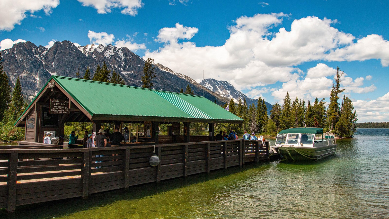 A real photograph of the Jenny Lake shuttle boat approaching the dock with mountains in the background and a small line of hikers waiting on the pier