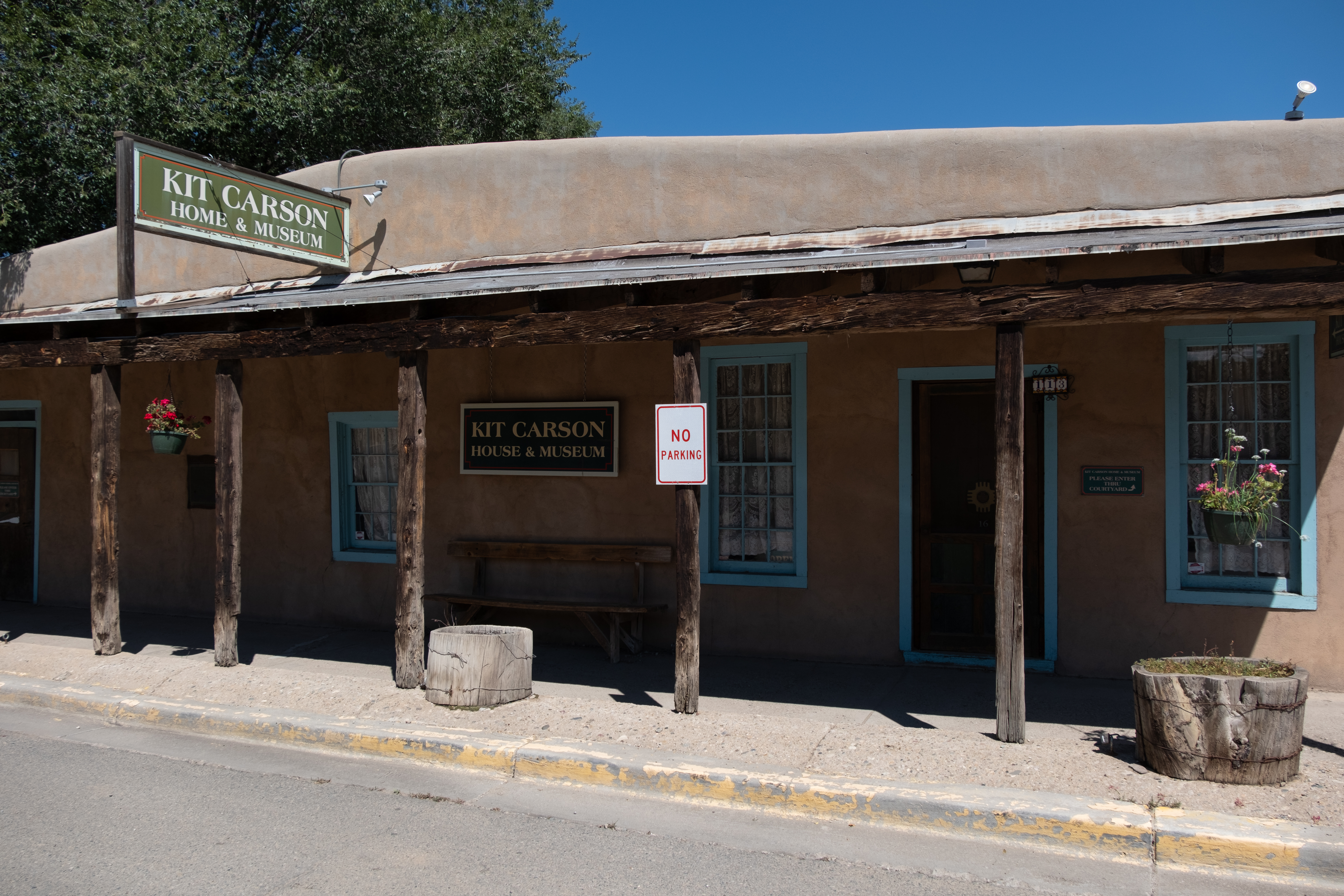 A real photograph of the Kit Carson Home and Museum in Taos, showing a low adobe exterior with a wooden door, a simple courtyard feel, and bright New Mexico sunlight