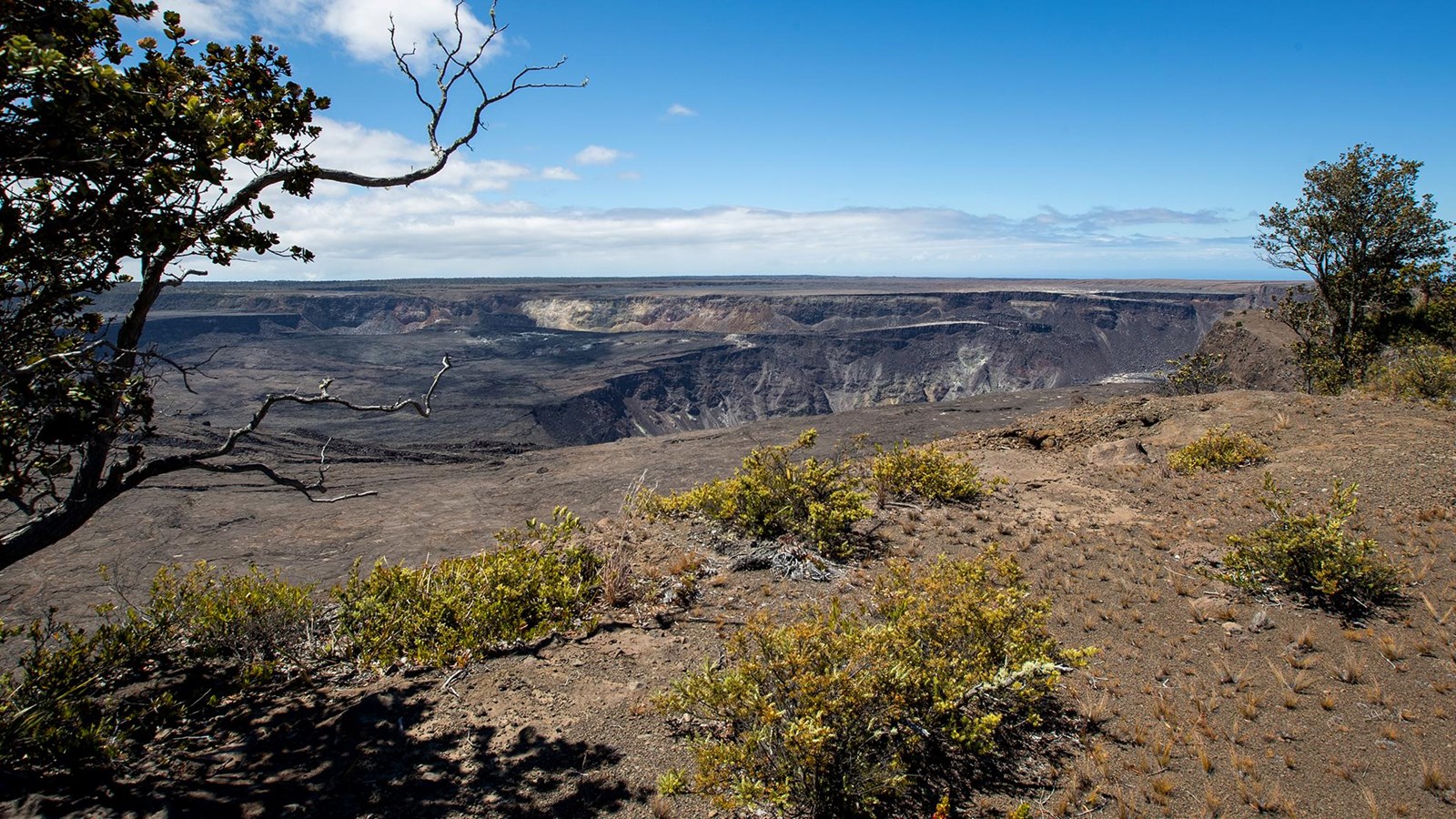 A real photograph of the Kīlauea caldera viewed from an overlook in Hawaiʻi Volcanoes National Park, with a wide volcanic crater, drifting steam, and dark lava rock under moody clouds