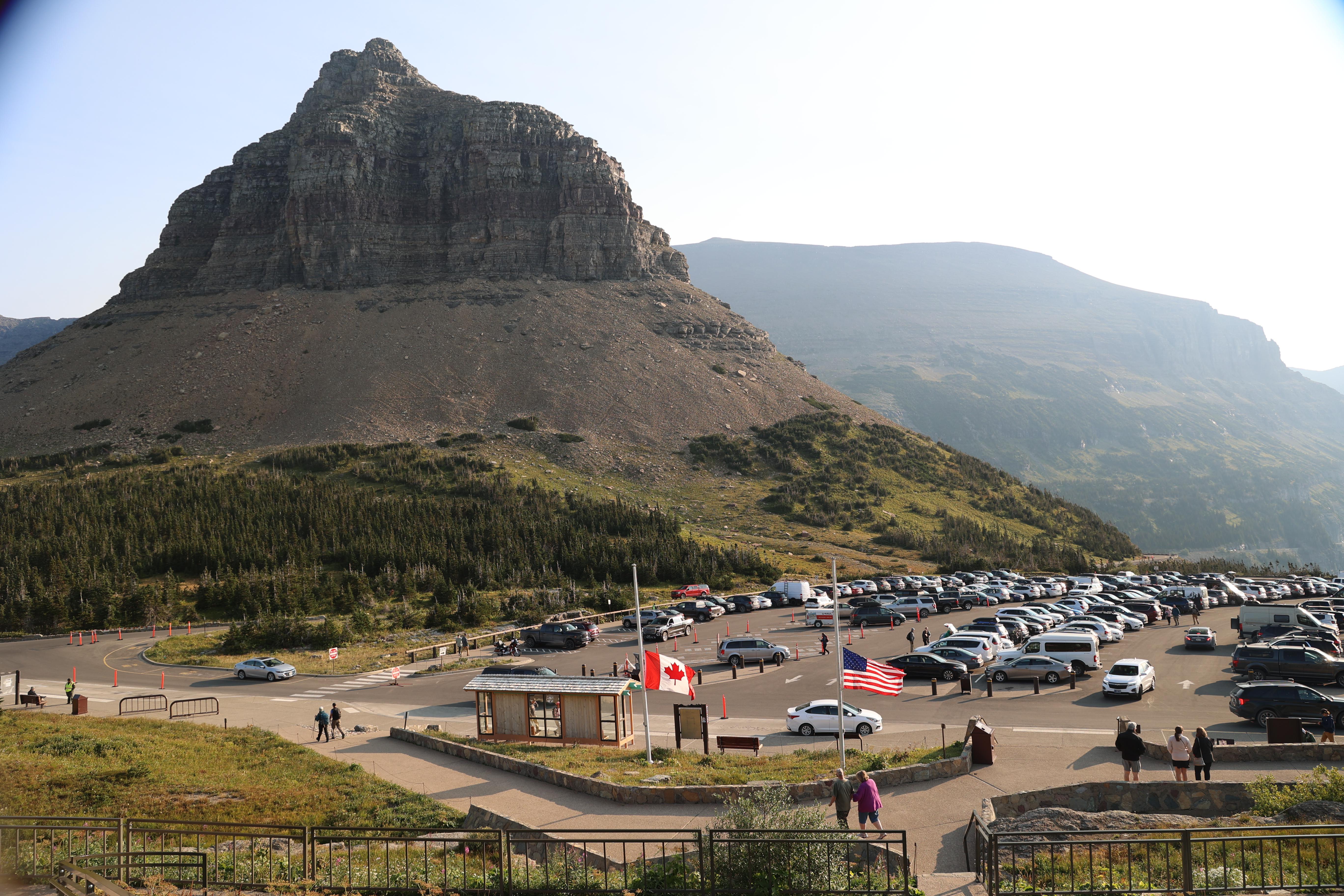 A real photograph of the Logan Pass visitor area in Glacier National Park in September, with cars parked along the lot and hikers in layered clothing walking toward the trailhead