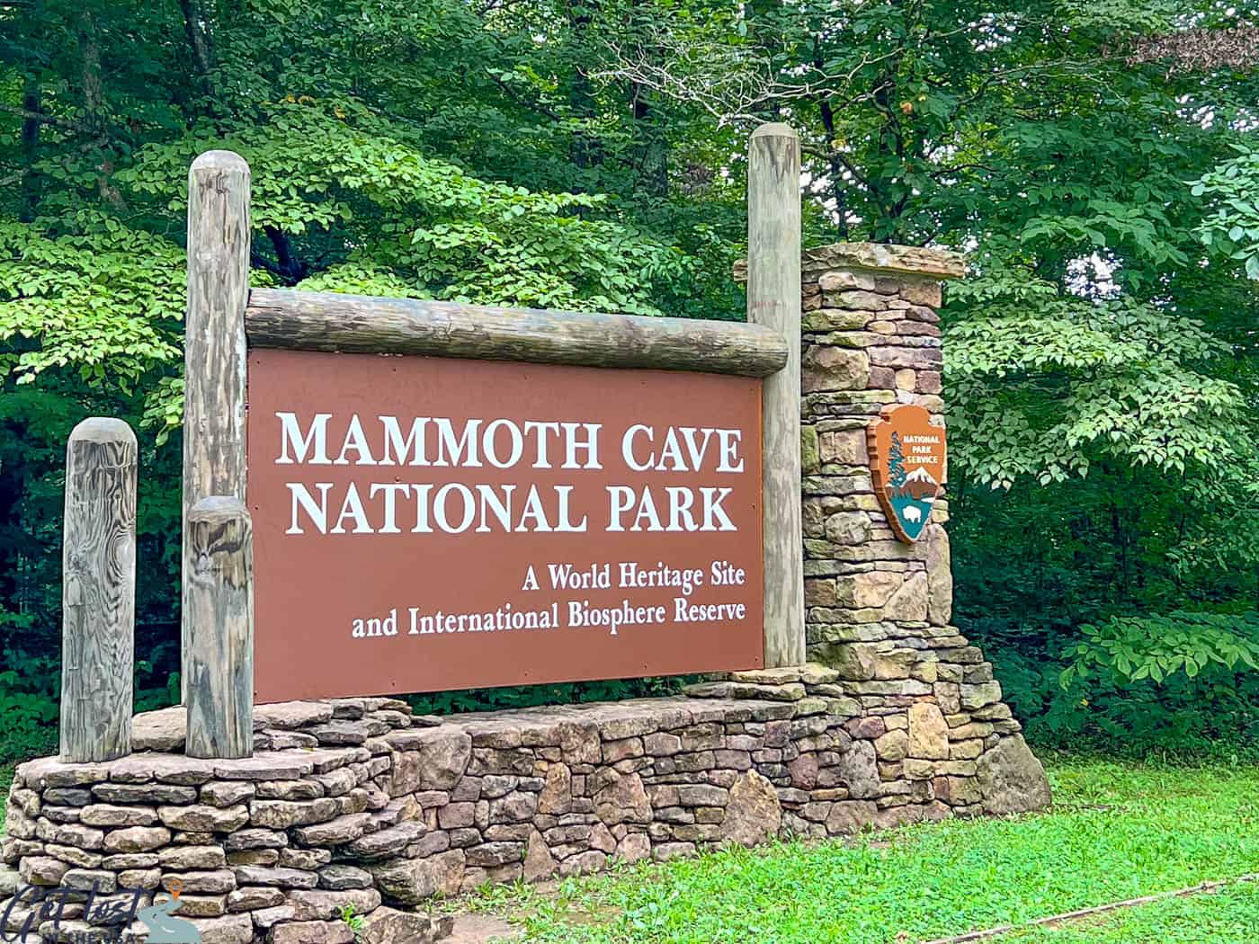 A real photograph of the Mammoth Cave Historic Entrance area in daylight, with stonework, signage, and visitors gathering near the opening surrounded by green forest