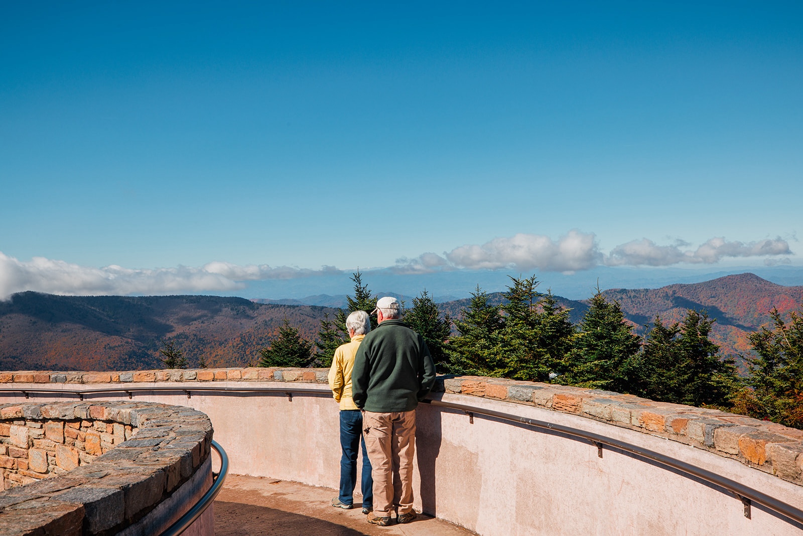 A real photograph of the Mount Mitchell summit area with the observation platform surrounded by evergreen trees, hikers in jackets, and hazy mountain layers beyond