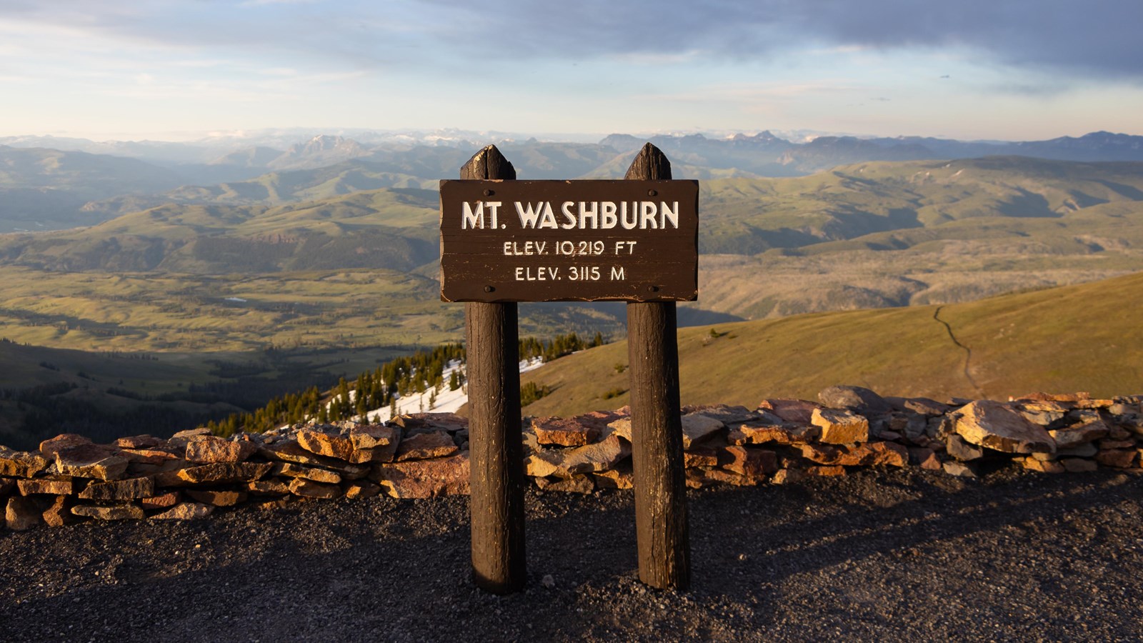A real photograph of the Mount Washburn summit area in Yellowstone with hikers near a lookout structure, rolling valleys below, and clouds casting shadows across the landscape