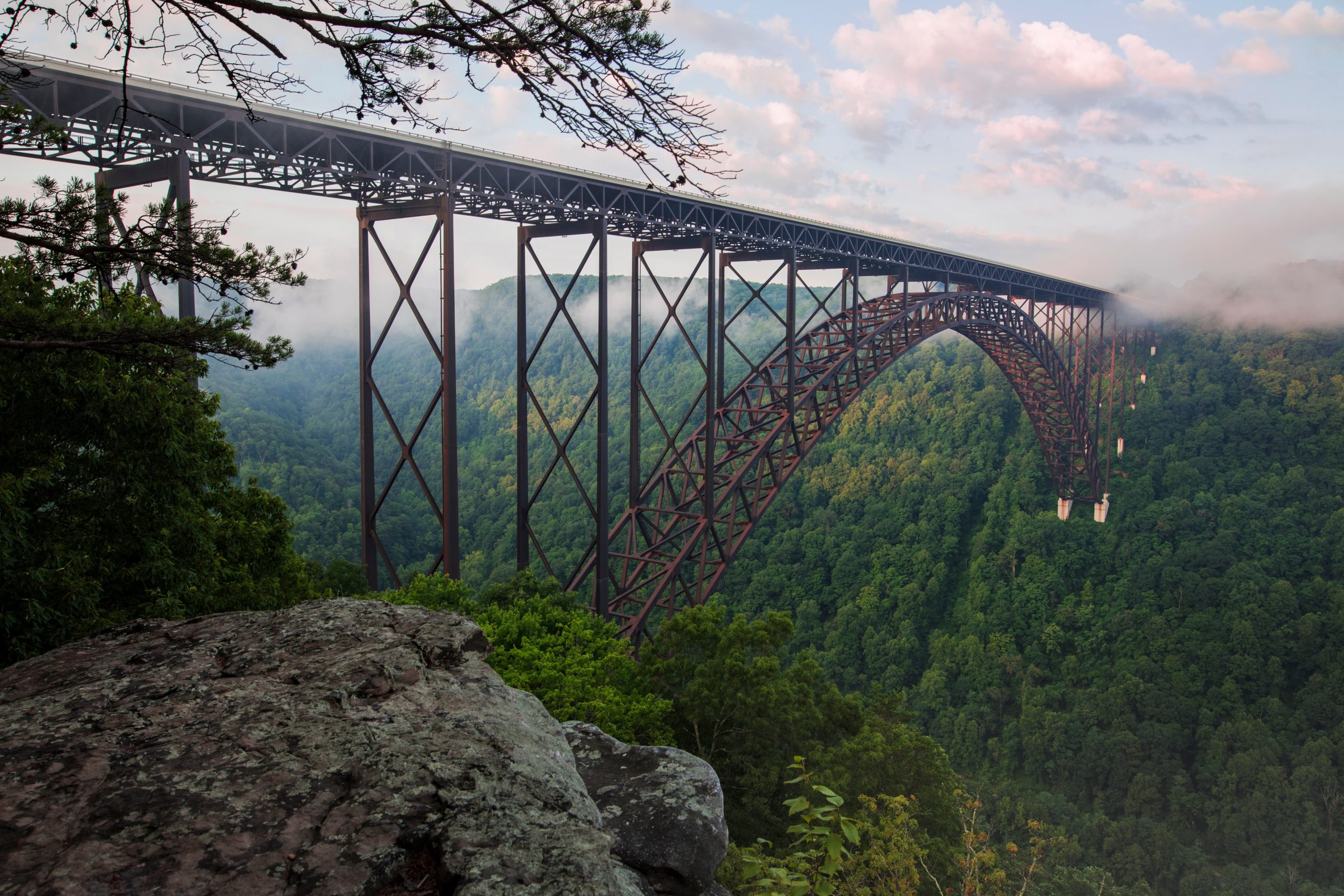 A real photograph of the New River Gorge Bridge spanning a forested canyon in West Virginia, seen from a popular overlook with morning light and layered ridgelines