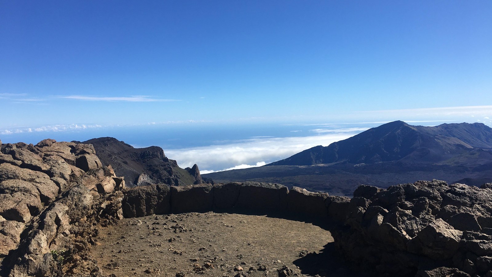 A real photograph of the Pā Kaʻoao (White Hill) Trail overlooking Haleakalā crater, with a cinder cone and layered clouds beyond
