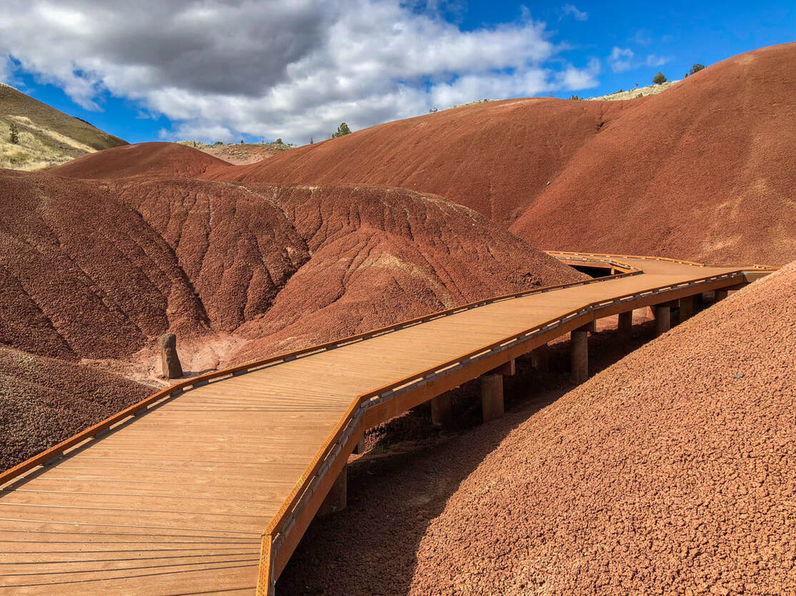 A real photograph of the Painted Cove Trail at Painted Hills in Oregon, showing a wooden boardwalk curving through red and tan clay hills on a clear day