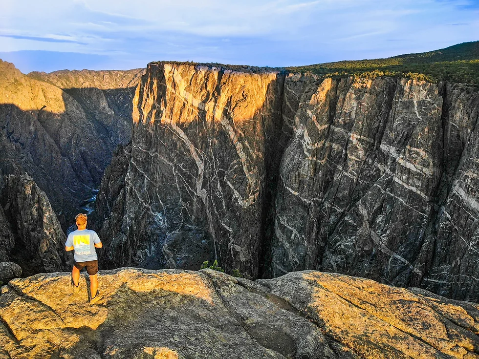 A real photograph of the Painted Wall view at Black Canyon of the Gunnison South Rim, with vertical dark rock cliffs dropping into a narrow canyon and layered ridgelines fading into the distance under clear Colorado light
