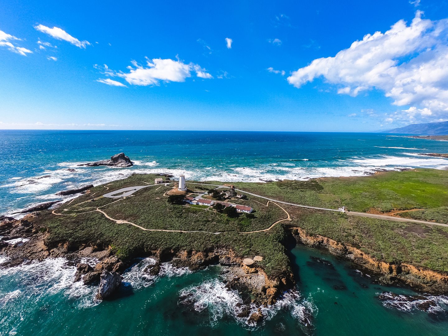A real photograph of the Piedras Blancas coastline near San Simeon with rocky shore, crashing waves, and the lighthouse visible in the distance under soft afternoon light