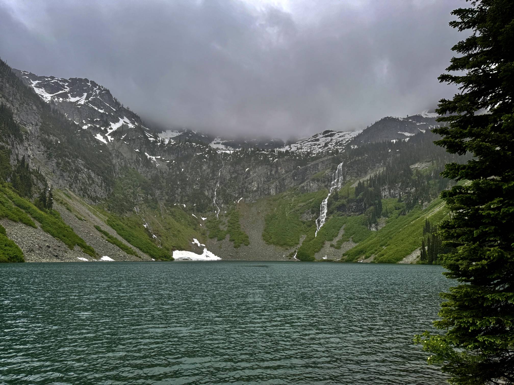 A real photograph of the Rainy Lake shoreline with calm water reflecting surrounding evergreen trees and distant rocky peaks on a bright summer day