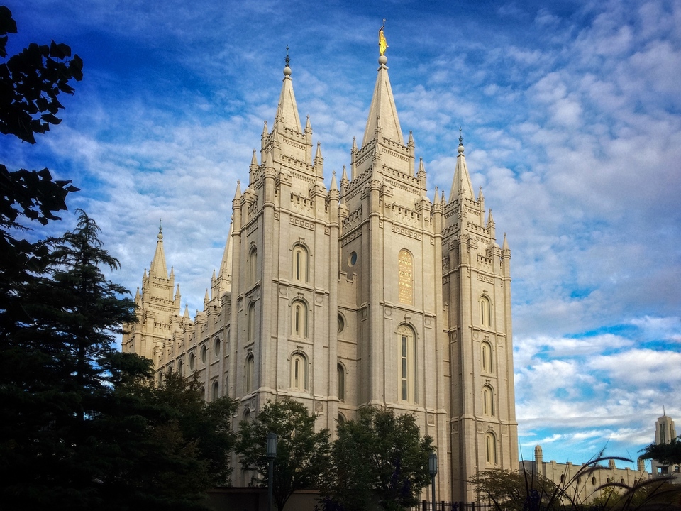 A real photograph of the Salt Lake Temple exterior seen from a pedestrian walkway, with manicured gardens and the Wasatch Mountains faintly visible beyond downtown