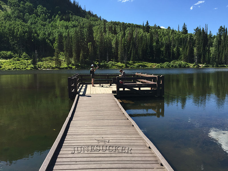 A real photograph of the Silver Lake trail near Brighton, Utah, showing a wooden boardwalk beside a calm mountain lake with evergreen trees and peaks in the background