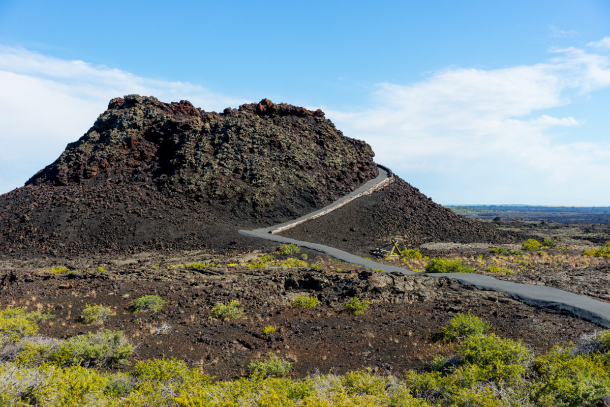 A real photograph of the Spatter Cones area at Craters of the Moon in Idaho, with a short path leading across dark lava to small rugged cones under clear daylight