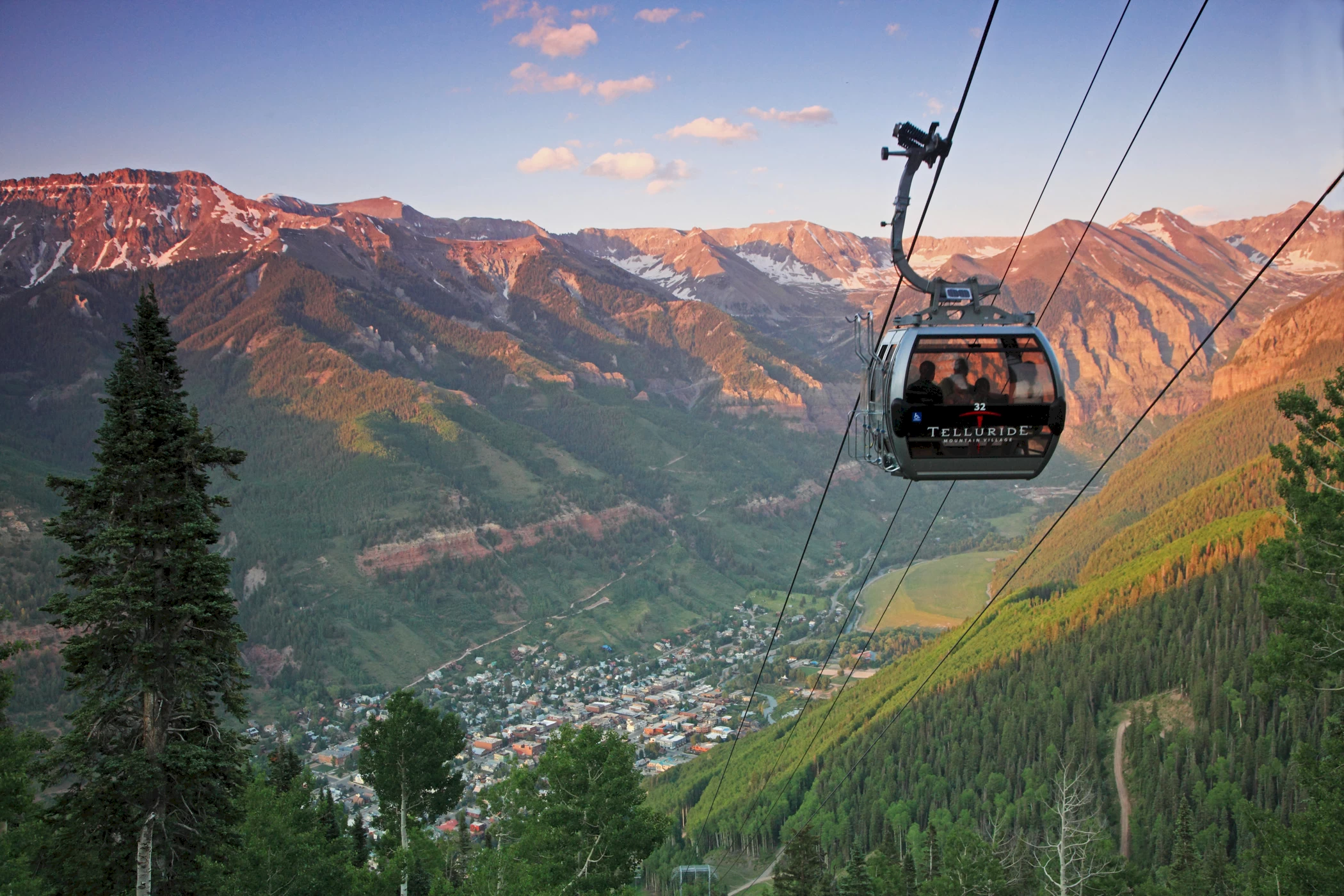 A real photograph of the Telluride gondola gliding above the valley in summer with green slopes and the town below, late afternoon light