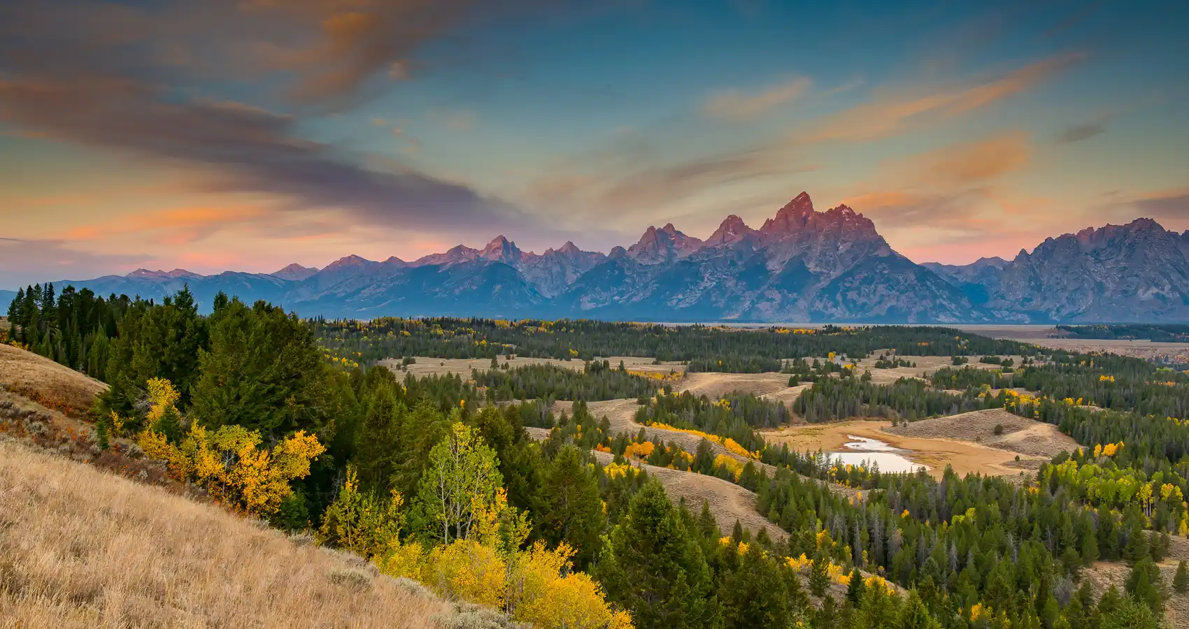 A real photograph of the Teton Range at sunrise from a valley viewpoint with pink alpenglow on jagged peaks and a calm foreground