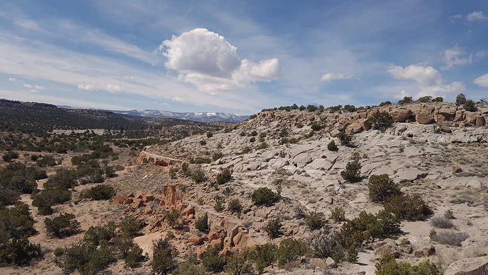 A real photograph of the Tsankawi loop trail on a wide mesa with tan volcanic rock, low vegetation, and distant mountains under a bright New Mexico sky