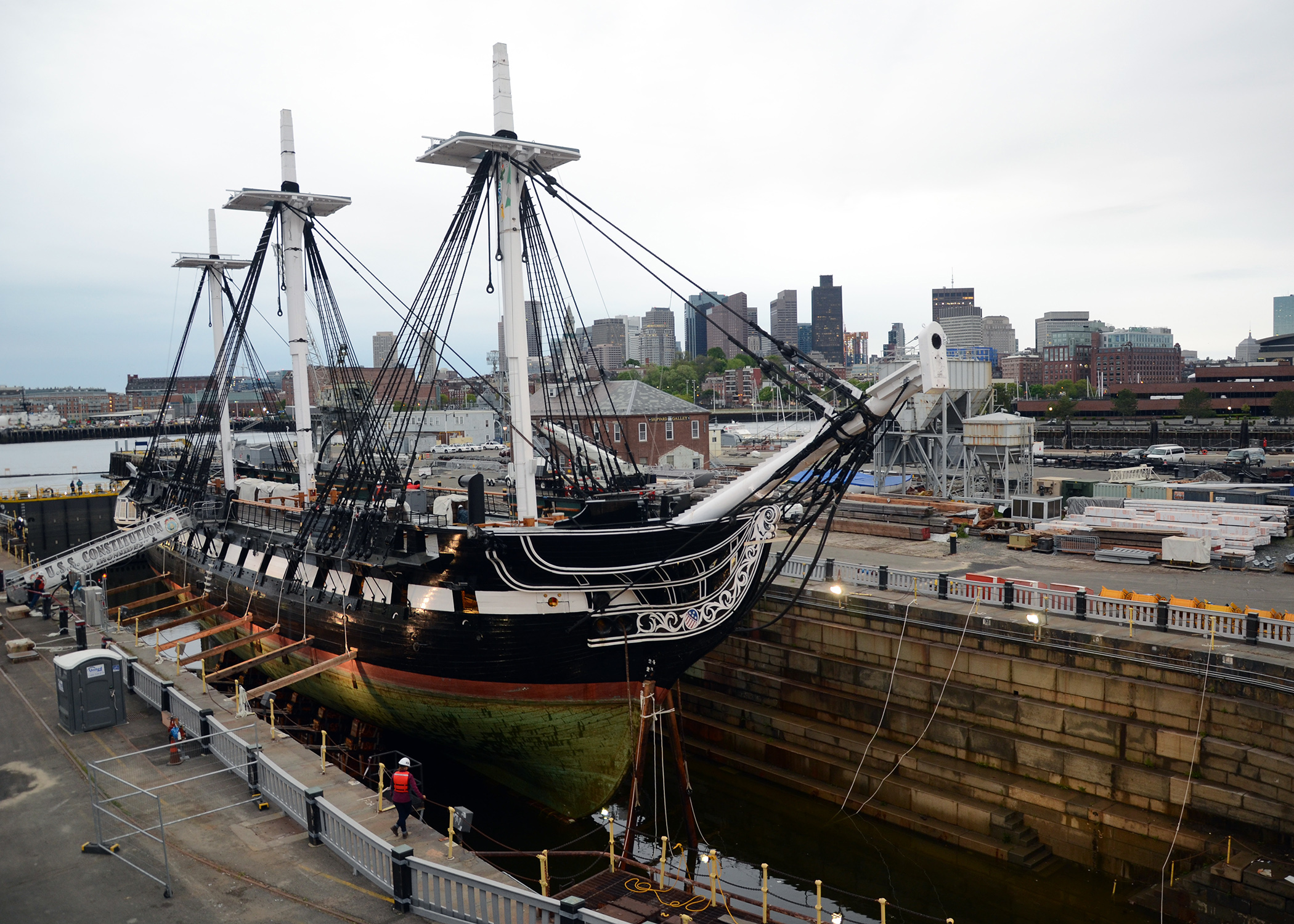 A real photograph of the USS Constitution docked at the Charlestown Navy Yard with rigging and masts against a bright sky