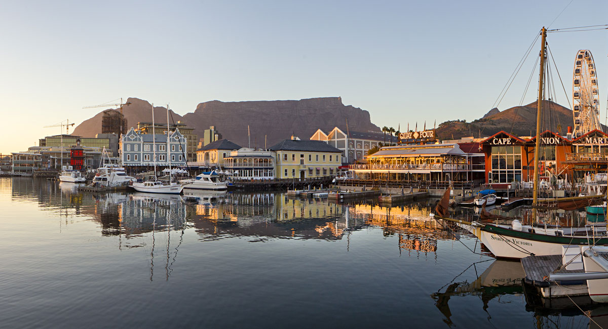 A real photograph of the V&A Waterfront in Cape Town at dusk, with restaurant lights reflecting on the harbor water and Table Mountain silhouetted in the background
