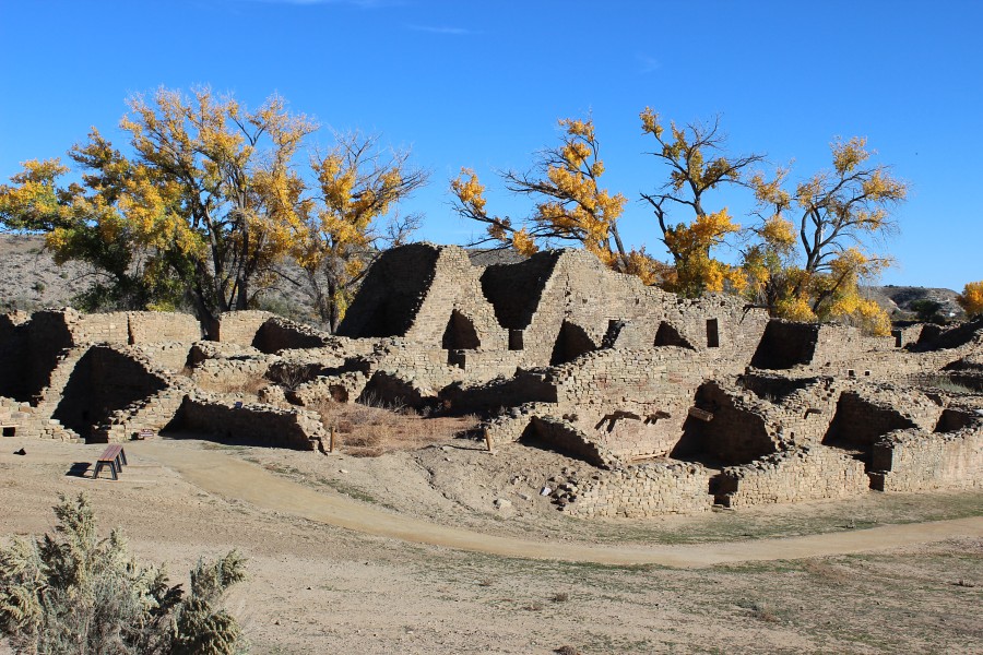 A real photograph of the West Ruins walking trail at Aztec Ruins National Monument, with a packed dirt path curving past low stone walls and ancient masonry under a bright New Mexico sky