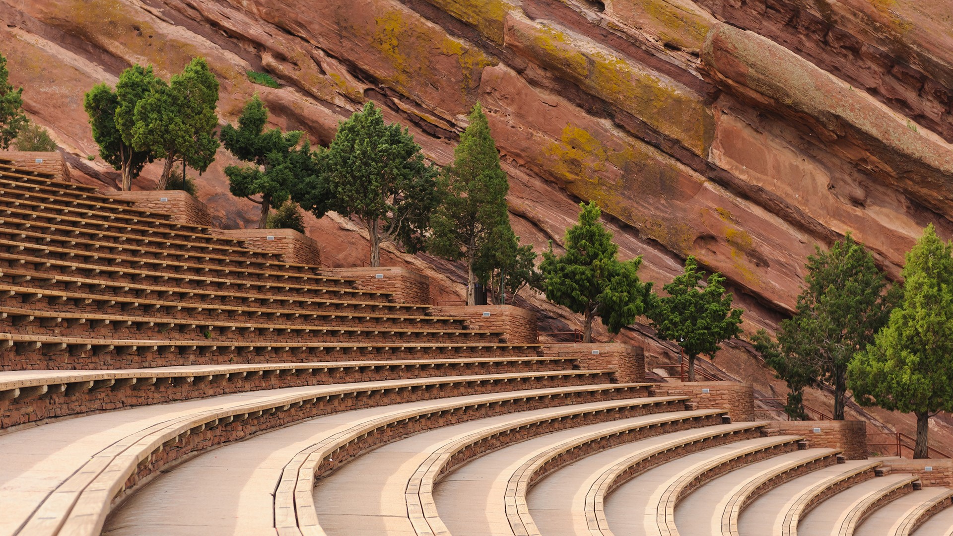 A real photograph of the empty seating rows at Red Rocks Amphitheatre in the morning, with red sandstone formations framing the view