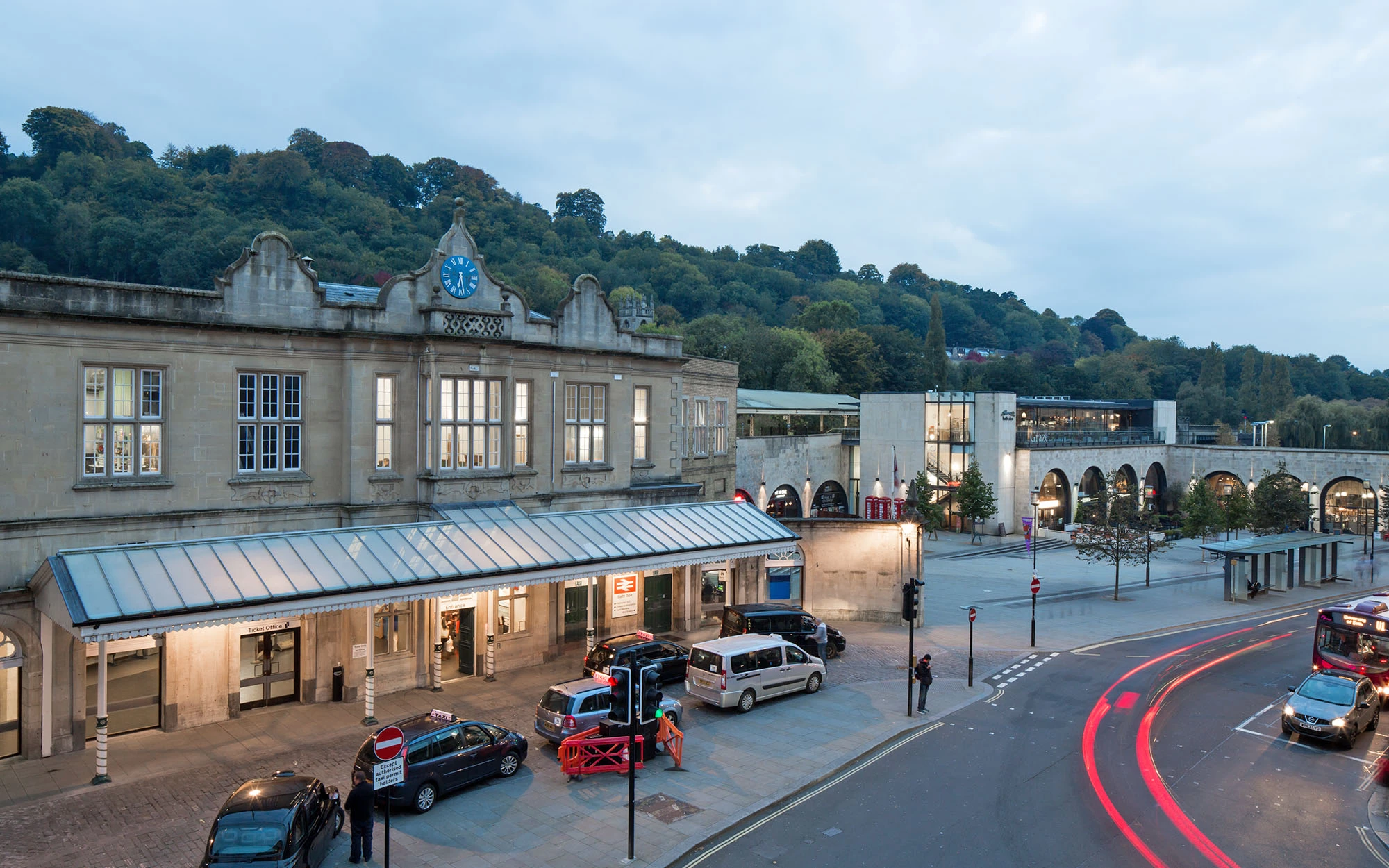 A real photograph of the exterior of Bath Spa railway station with travelers walking out toward the city, buses and taxis nearby, overcast soft light, candid travel photography