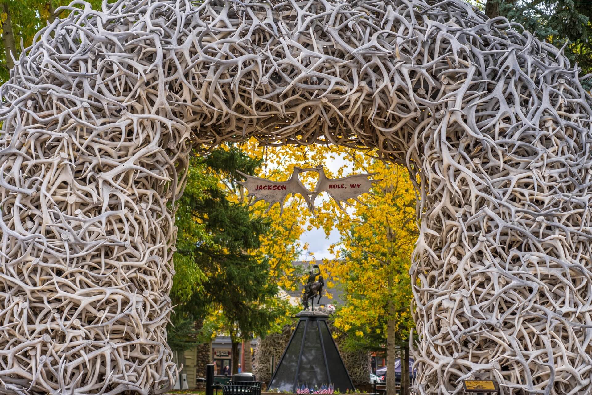 A real photograph of the iconic elk antler arches at Jackson Town Square on a crisp early fall morning, with warm sunrise light and a few people walking through the square