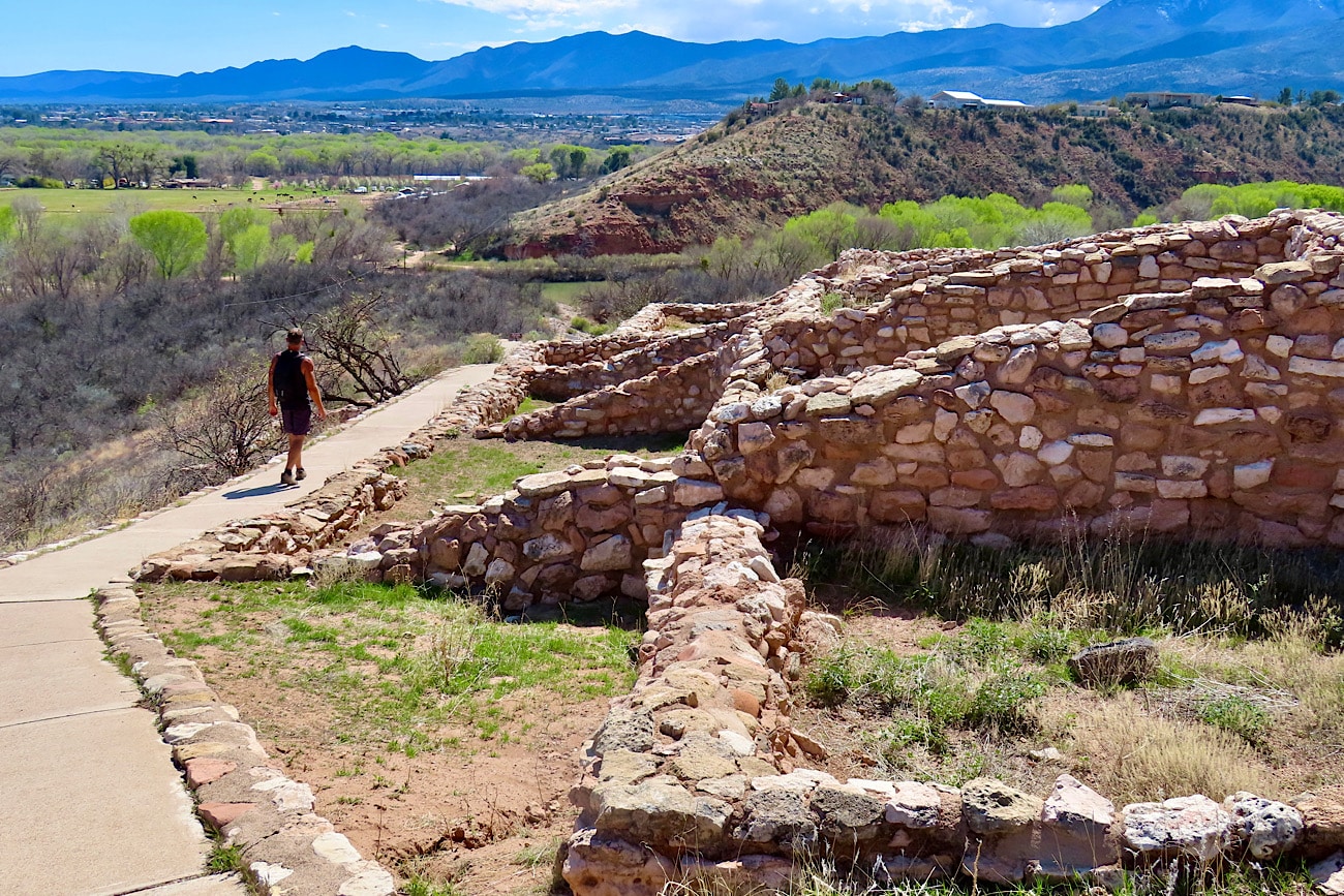 A real photograph of the paved Pueblo Trail path curving past low stone walls at Tuzigoot with desert shrubs and bright midday light