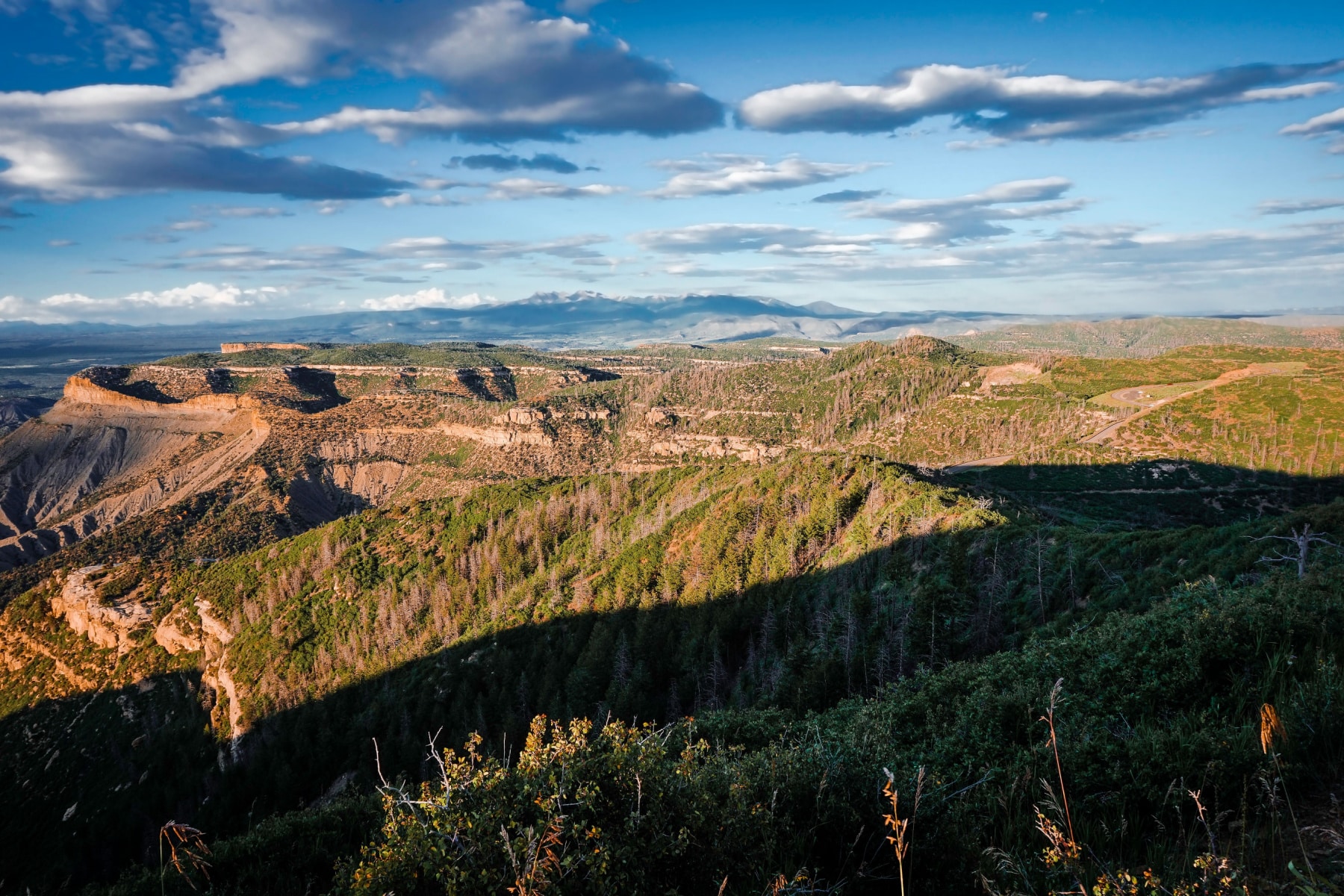 A real photograph of the sun setting over a broad canyon at Mesa Verde National Park, with layered mesas fading into the distance and warm golden light across the landscape