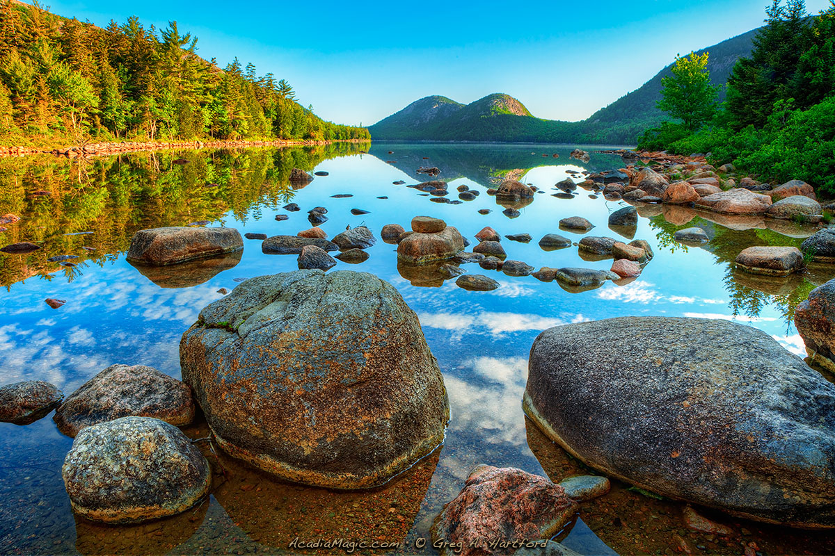 A real photograph of the wooden shoreline path at Jordan Pond in Acadia National Park on a calm morning, with clear water reflecting pine trees and the Bubbles mountains in the distance