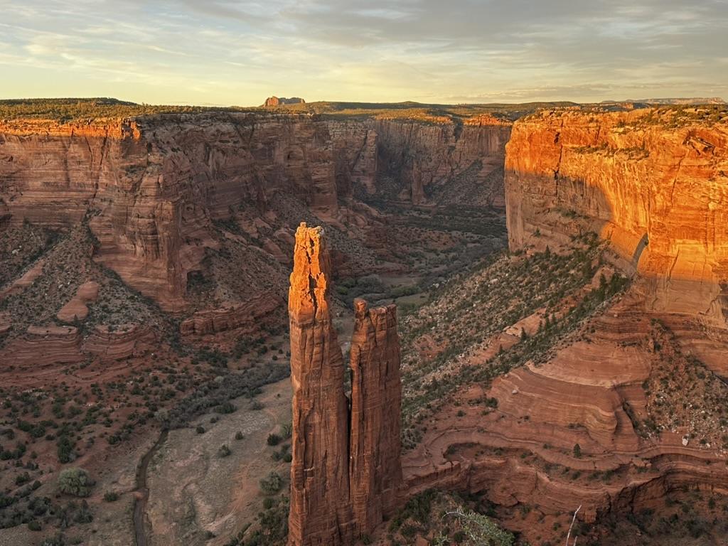 A real photograph of two visitors standing at a Canyon de Chelly South Rim overlook near sunset, looking out over red canyon walls under warm golden light