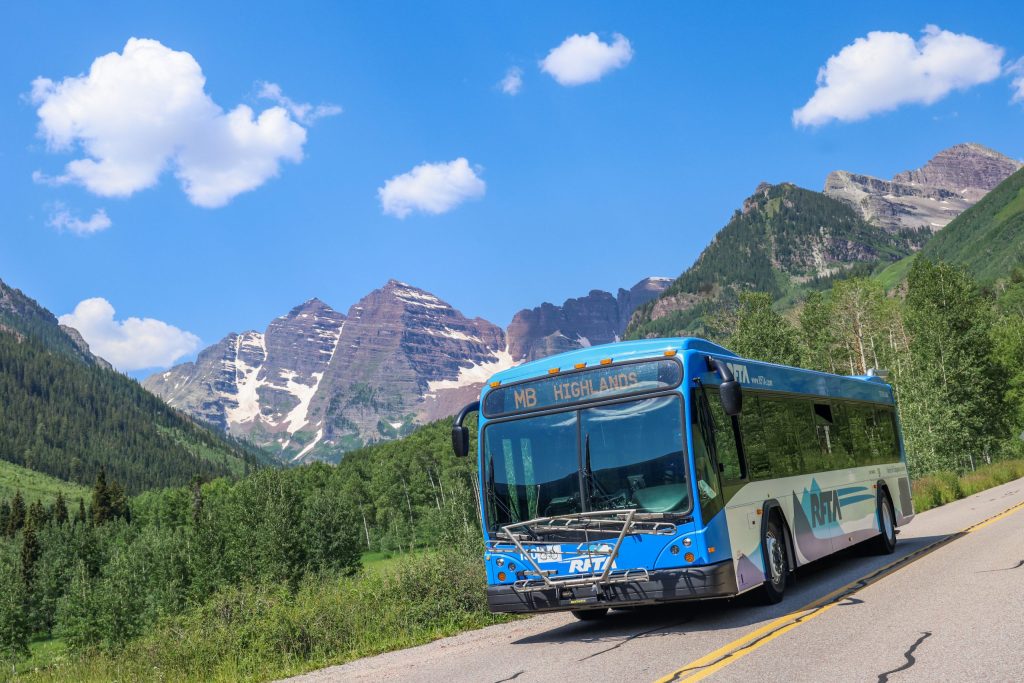 A real photograph of visitors lining up at the Maroon Bells shuttle boarding area at Aspen Highlands with buses and mountain scenery in the background