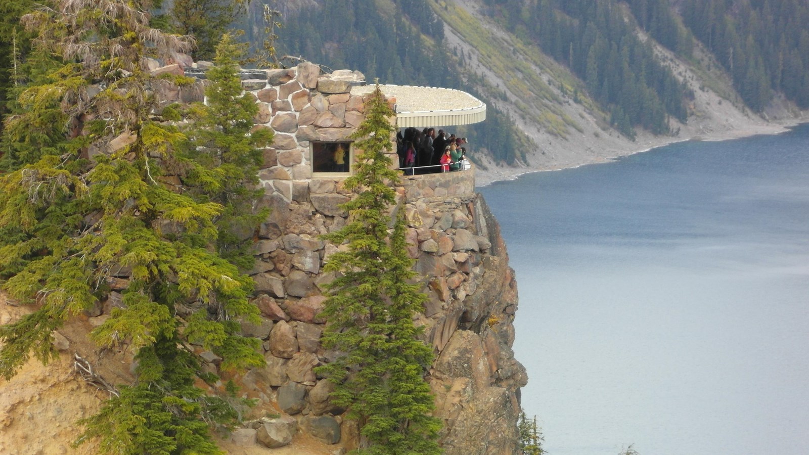 A real photograph of visitors standing at a stone overlook wall at Rim Village with Crater Lake’s deep blue water filling the background
