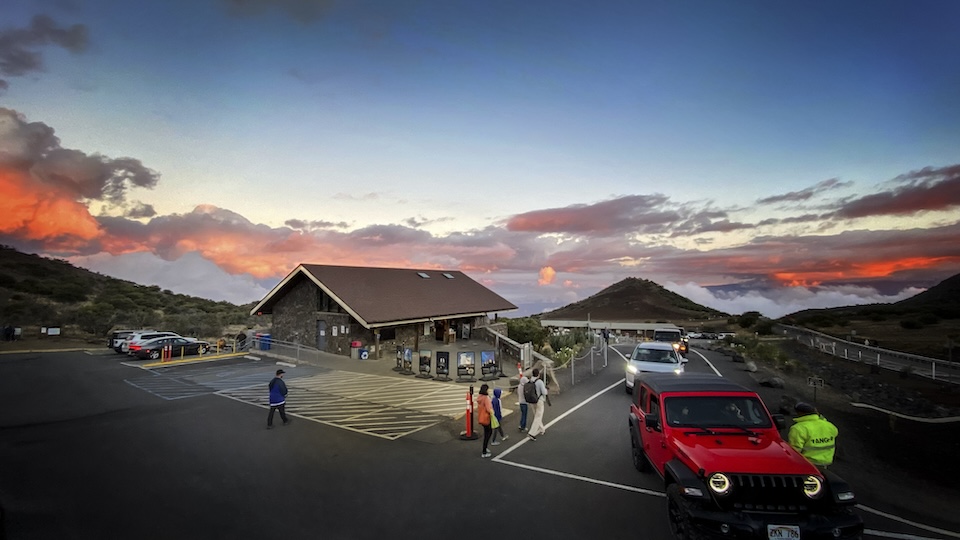 A real photograph of visitors standing near the Mauna Kea Visitor Information Station at sunset, with the sun low over the cloud layer and the volcanic landscape in the foreground