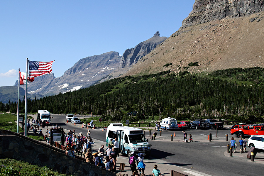 A real photograph of visitors waiting at a shuttle bus stop near Logan Pass in Glacier National Park, with mountains in the background and people wearing daypacks and outdoor layers