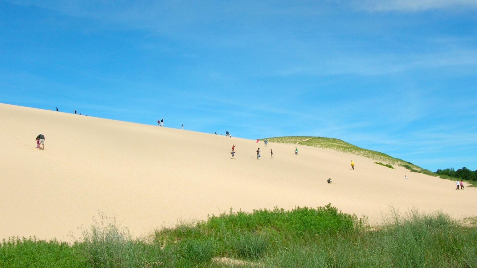 A real photograph of visitors walking up the steep sandy slope at the Sleeping Bear Dunes Dune Climb with footprints in the sand and Lake Michigan visible in the distance