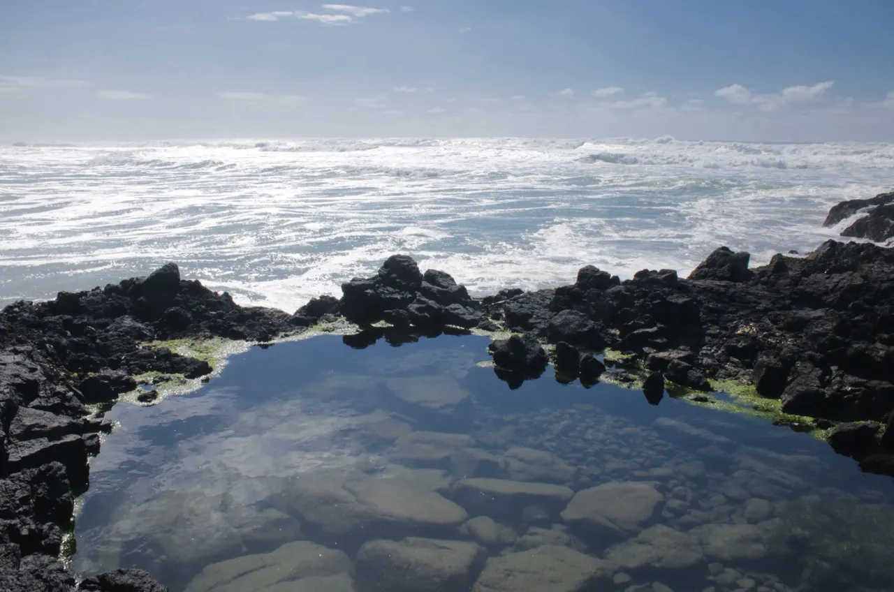 A real photograph of waves crashing into Devil's Churn at Cape Perpetua with white spray rising from dark basalt cliffs under a gray sky
