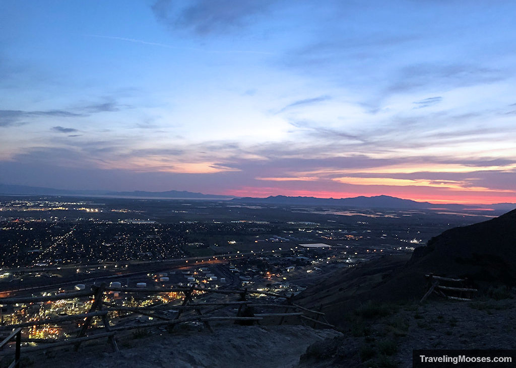 A real photograph taken at sunset from Ensign Peak overlooking Salt Lake City, with the downtown grid below and the Wasatch Mountains silhouetted in warm evening light