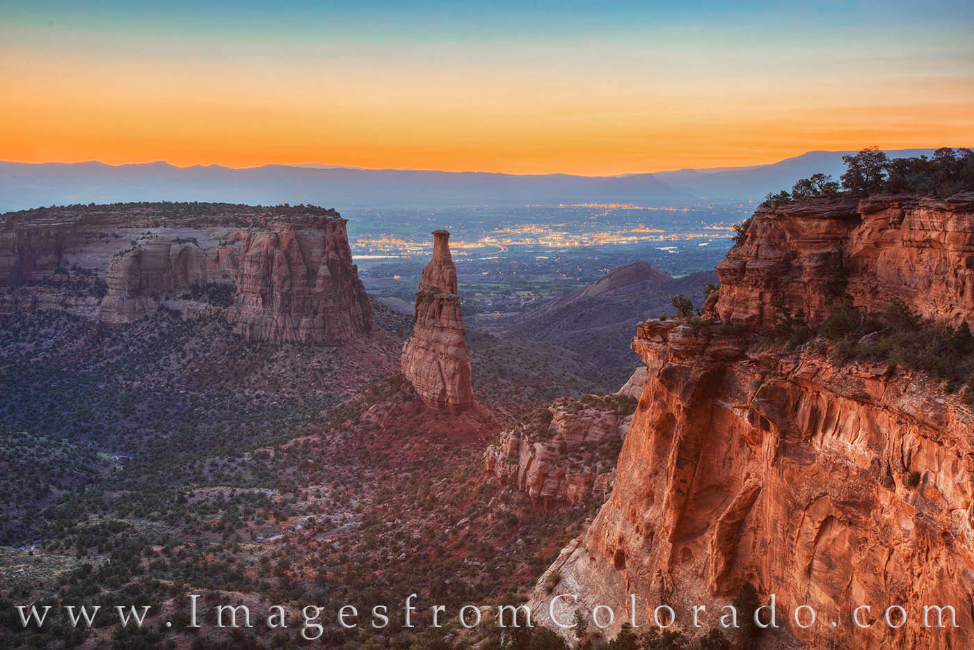 A real photograph taken at sunset from a Rim Rock Drive overlook in Colorado National Monument, with glowing red sandstone cliffs dropping into a canyon and distant mesas under a clear sky