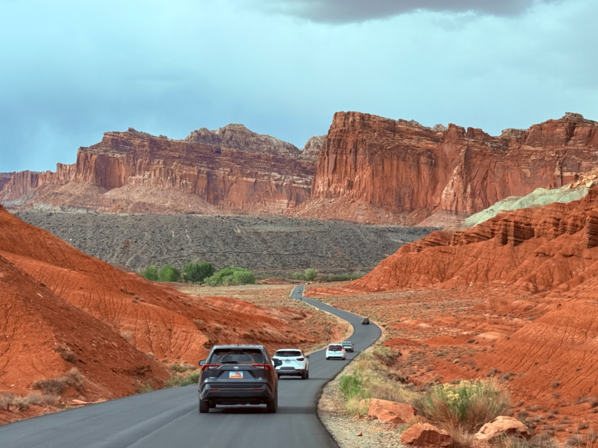 A real photograph taken from an overlook along Capitol Reef Scenic Drive showing a paved road winding through red rock cliffs and desert vegetation under a clear blue sky