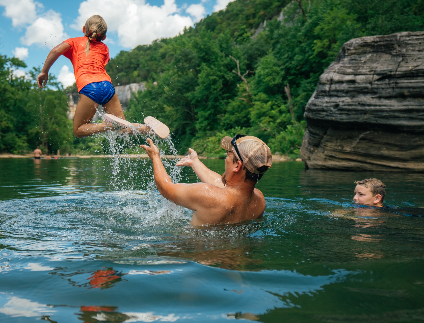 A real summer photograph of people wading and swimming near a wide gravel bar on the Buffalo National River, with tall limestone bluffs and green forest in the background