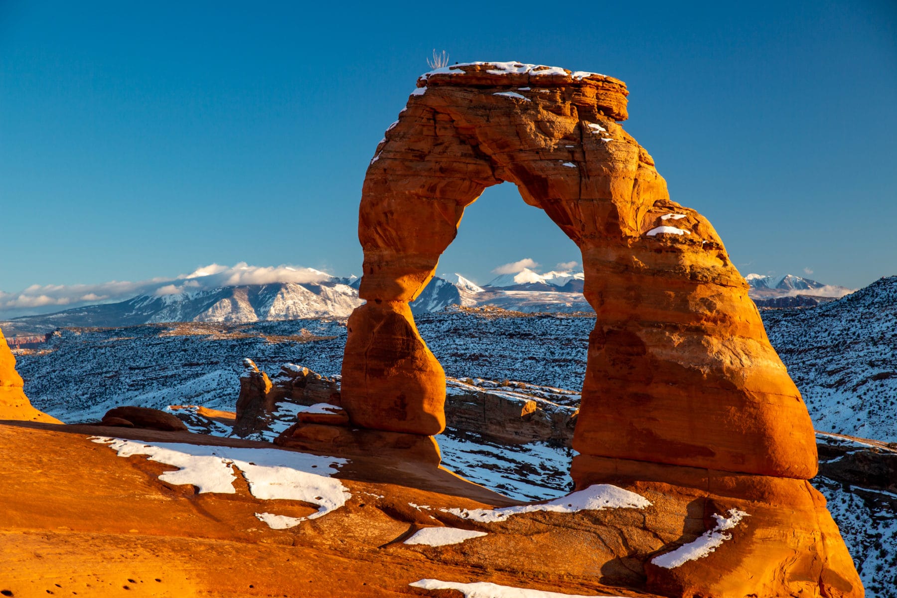 A real winter photograph of Delicate Arch in Arches National Park with a light dusting of snow on the red sandstone and the La Sal Mountains visible in the distance under clear blue sky