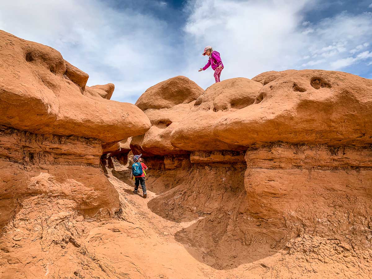 A realistic photograph of a parent holding a child’s hand while walking on smooth slickrock near sandstone hoodoos in Goblin Valley, showing careful footing and desert sunlight