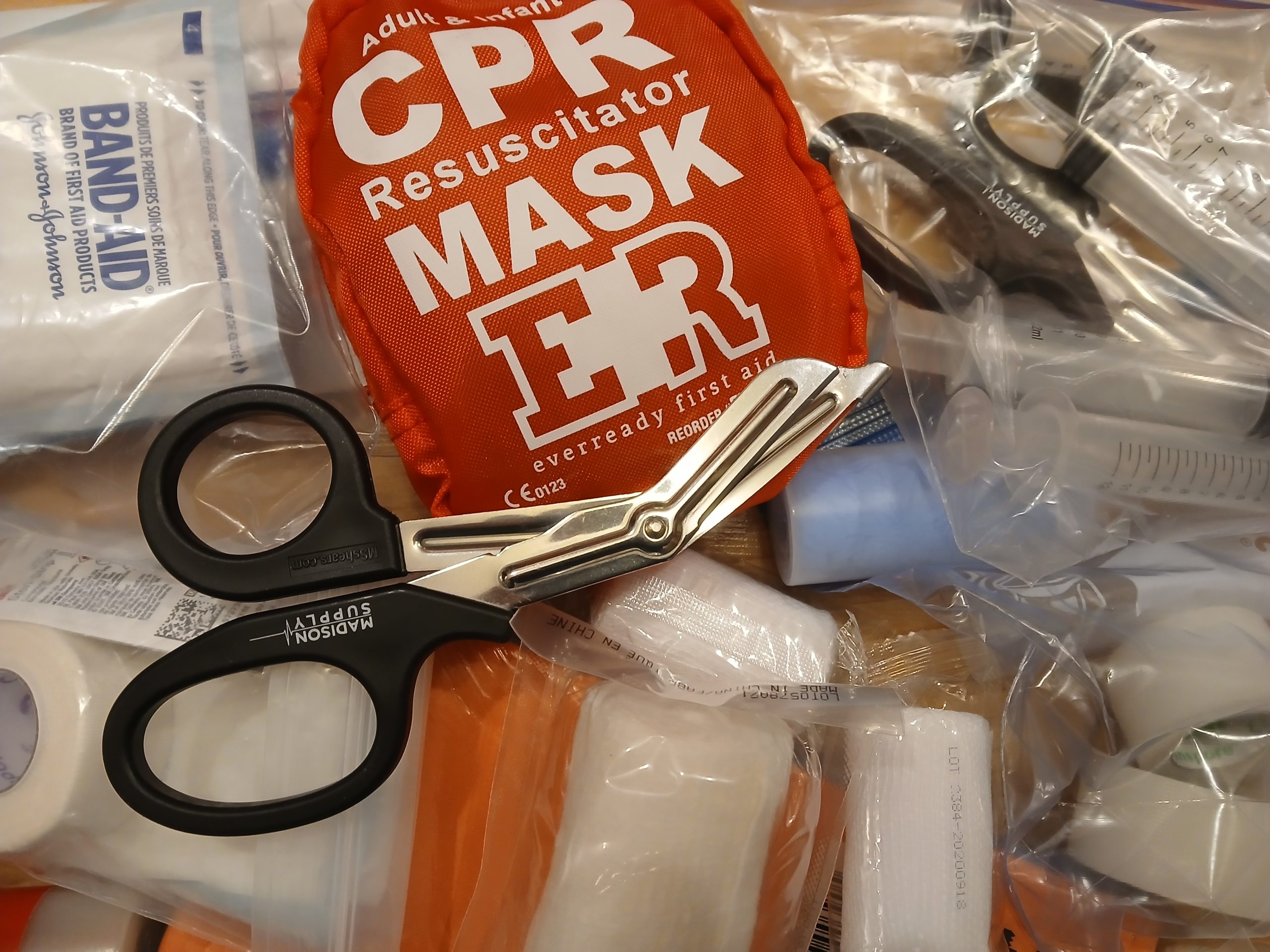 A realistic photograph of a small hiking first aid pouch open on a wooden picnic table with fine-tipped tweezers, alcohol wipes, and a small bottle of insect repellent visible