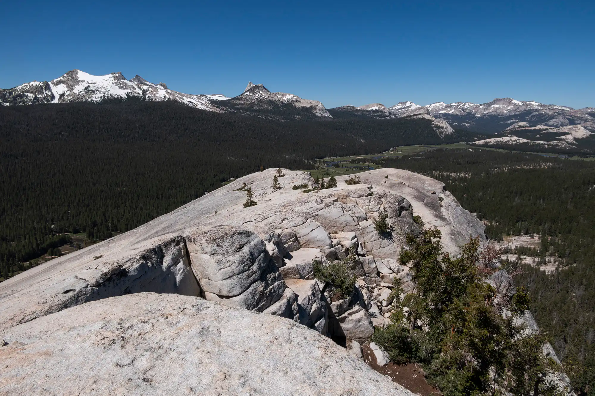 A realistic summit-view photograph from Lembert Dome showing Tuolumne Meadows below, scattered pines, and granite peaks under a clear afternoon sky
